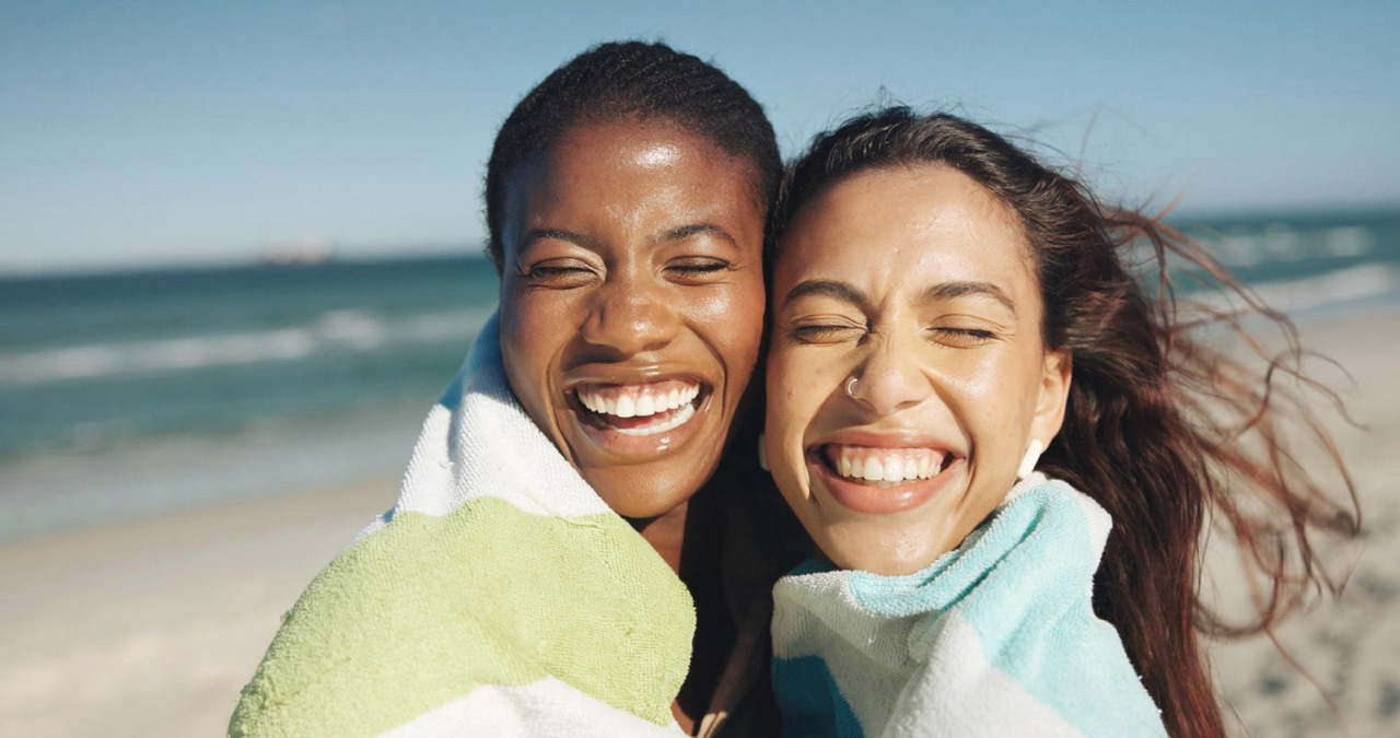 Two women laughing on the beach wrapped in towels representing summer skincare routine for hormonal acne