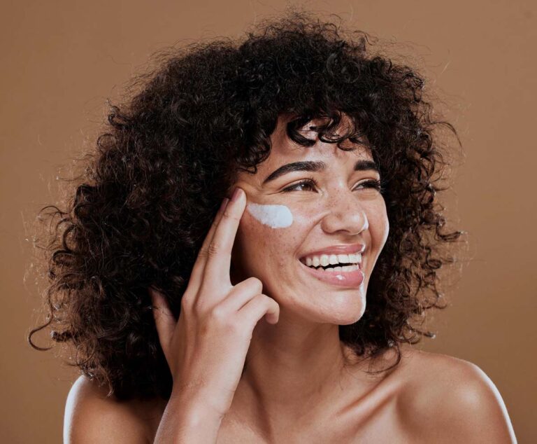 Smiling woman applying face cream to her cheek, deciding between sunscreen and moisturizer